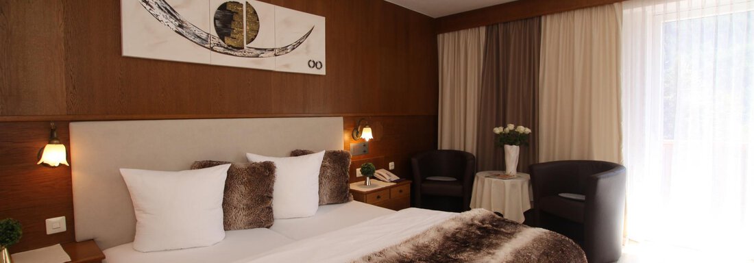A modern bedroom with a large bed and stylish furniture. The walls are in warm wood tones, and the window lets in plenty of daylight. - Haus Amaris - Sölden
