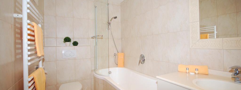 A modern bathroom with a bathtub and a shower. The walls are light tiled, and orange towels are decoratively placed. - Haus Amaris - Sölden