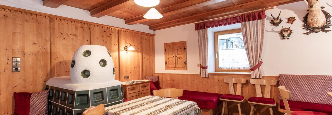 A cozy room with wooden paneling and a traditional bench. In the center stands a tiled stove, and there is a table with chairs. - Haus Bergblick - Sölden
