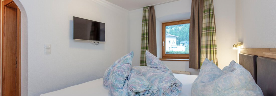 A cozy bedroom with a double bed and two large pillows. Next to the bed is a television and a window with curtains. - Haus Bergblick - Sölden