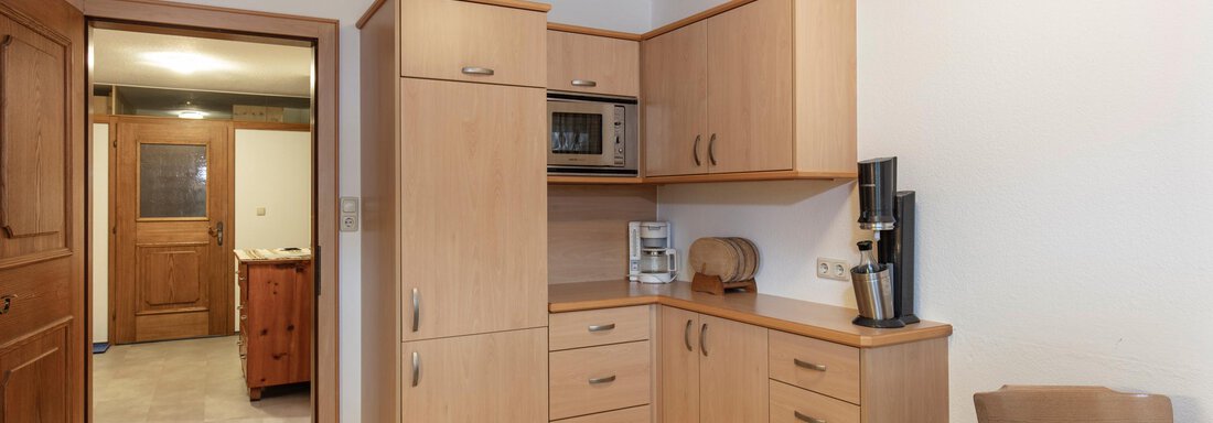 A modern kitchen with wooden cabinets and appliances. A table is visible in the foreground, while the door to another room is open in the background. - Haus Bergblick - Sölden
