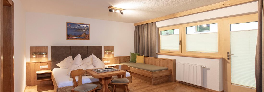 A cozy room with wooden decor, a double bed, and a table with chairs. Large windows provide plenty of daylight and a welcoming atmosphere. - Haus Bergblick - Sölden