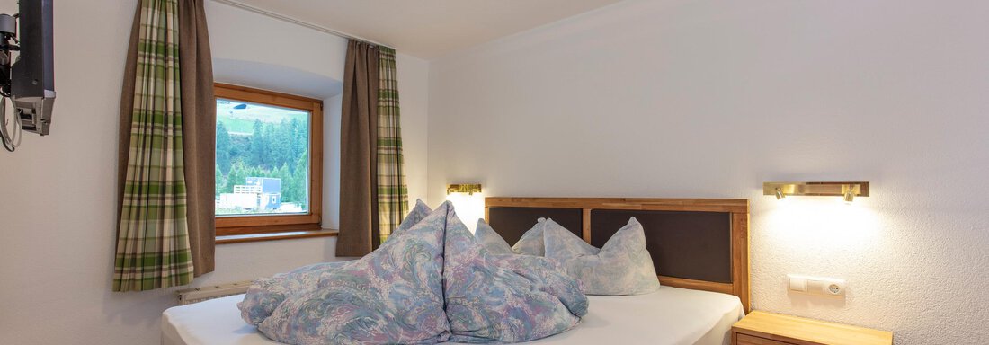 A cozy bedroom with a large bed and colorful cushions. There is a window with a view of greenery and a television on the wall. - Haus Bergblick - Sölden