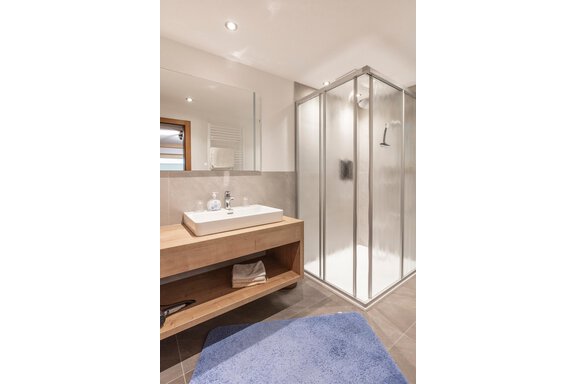 A modern bathroom with a spacious shower. The sink area is made of wood and there is a blue rug on the floor. - Haus Bergblick - Sölden