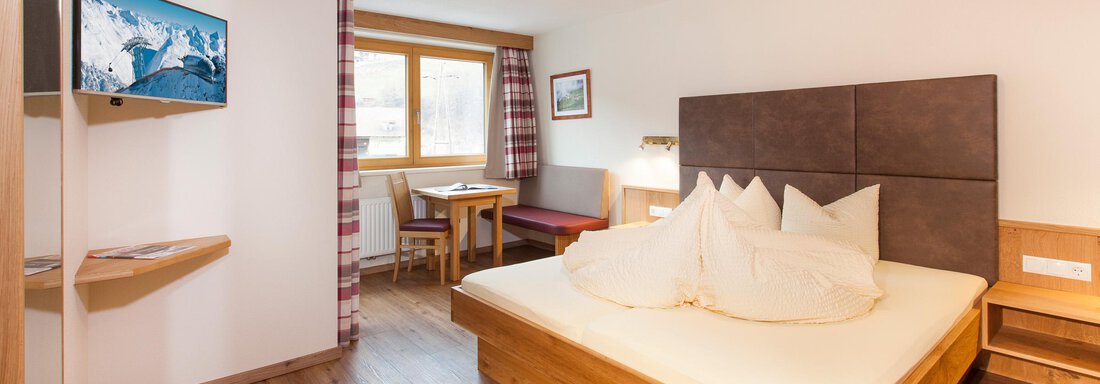 Doppelzimmer A cozy hotel room with a large bed, a television, and a dining table. The room has large windows and a warm wooden decoration. - Haus Bergblick - Sölden