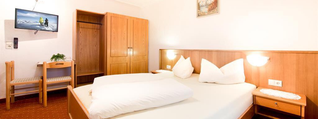 A cozy room with a double bed and two pillows. There is a wooden dresser, a desk, and a television on the wall. - Haus Christian - Sölden