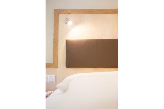 A cozy bedroom with a light wood frame and a brown headboard. The bed is covered with a white blanket and there is a wall lamp. - Haus Christian - Sölden