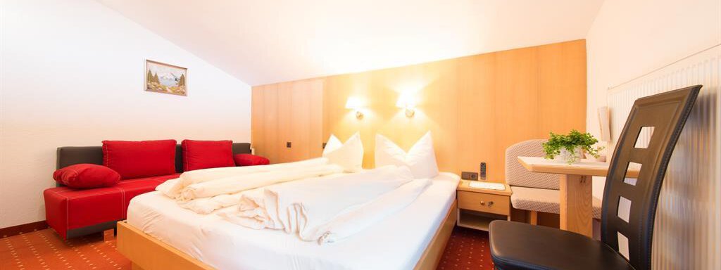 A cozy hotel room with a double bed and a red sofa. There is a small table and a chair, as well as soft lighting. - Haus Christian - Sölden