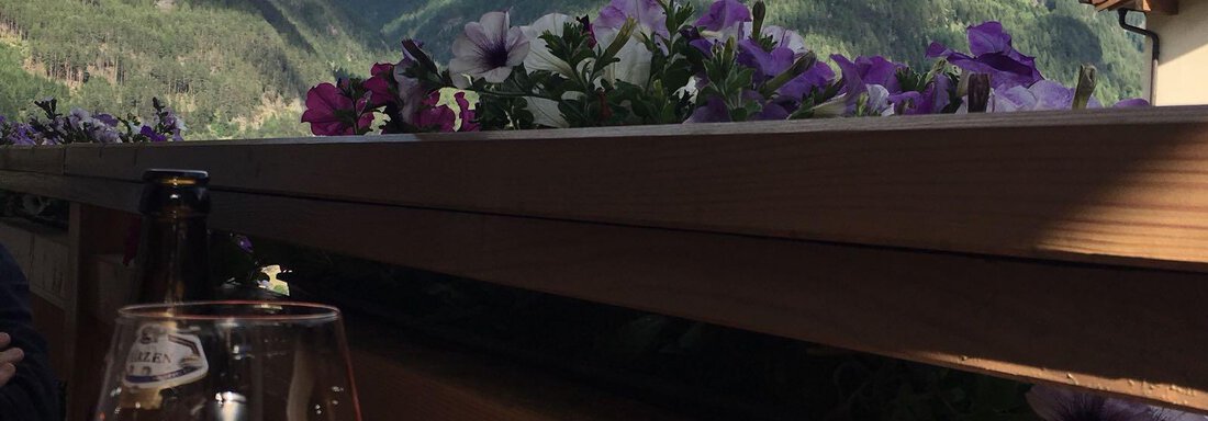 A glass of pink drink stands on a table with flowers in the foreground. In the background, there are tall mountains and a blue sky with clouds. - Haus Edmund - Sölden