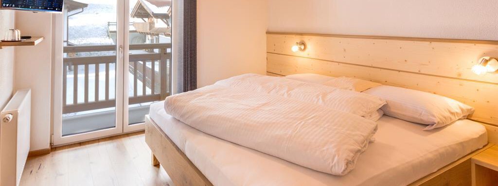 A cozy bedroom with a large bed and wooden details. There is a TV on the wall and a balcony with an outdoor view. - Haus Edmund - Sölden