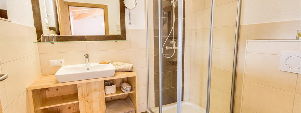 A modern bathroom with a shower and a wooden vanity. The walls are bright and the floor is designed with dark tiles. - Haus Edmund - Sölden