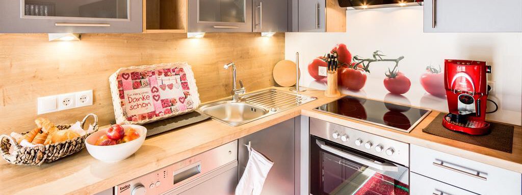 A modern kitchen with gray cabinets and wooden surfaces. There is a stove, a sink, and a decorative wall with tomato motifs. - Haus Edmund - Sölden