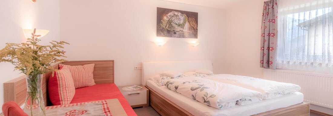 A cozy bedroom with a double bed and a red sofa bed. The room is bright and friendly, with a large window and modern decor. - Haus Edmund - Sölden