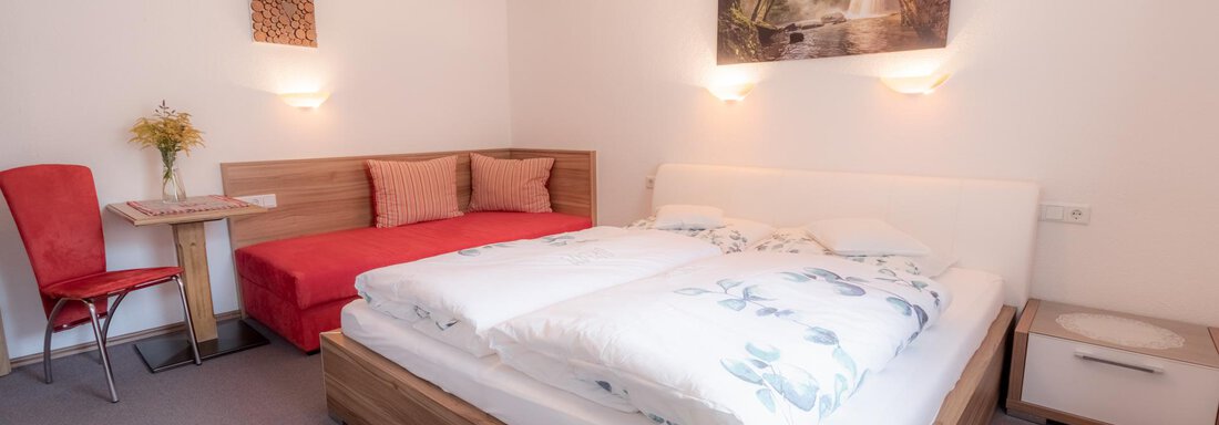 A cozy room with a large bed and a red sofa. The walls are light and there is a modern piece of art above the bed. - Haus Edmund - Sölden