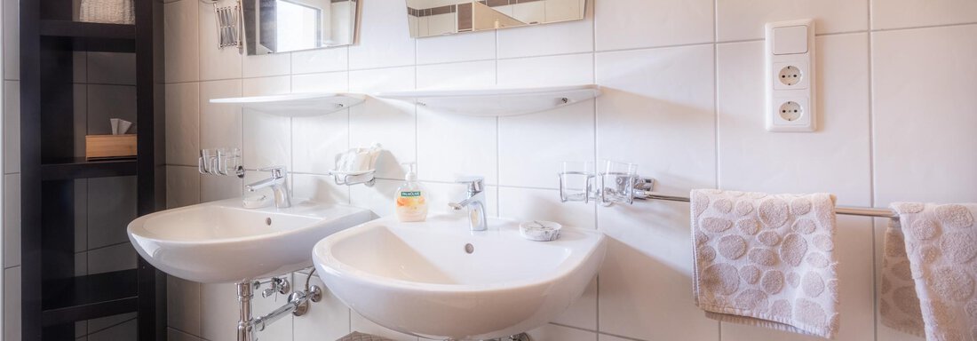 A modern bathroom with two sinks and large mirrors. The walls are tiled and there is a black shelving unit for storage. - Haus Edmund - Sölden