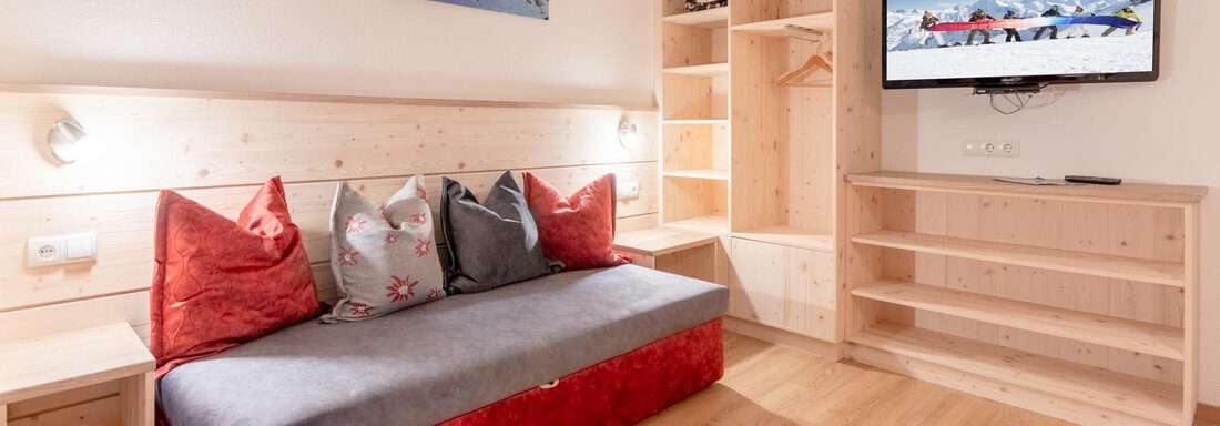A cozy living space with a red sofa and decorative pillows. A television hangs on the wall and there is a wooden shelf wall. - Haus Edmund - Sölden