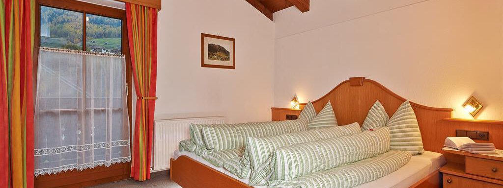 Schlafzimmer A cozy bedroom with a large bed and a wooden ceiling. Next to the bed is a nightstand and a window with curtains. - Haus Fontana - Sölden