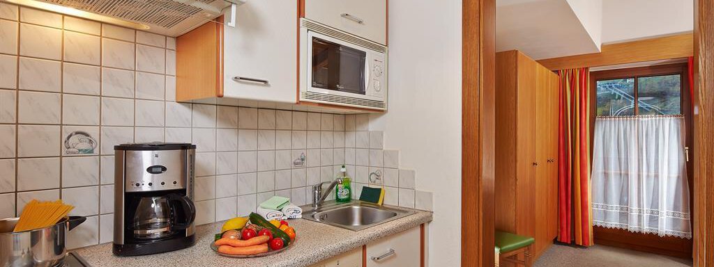 A cozy kitchen with modern appliances and a bright workspace. In the background, there is a passage to another room with a window and curtains visible. - Haus Fontana - Sölden
