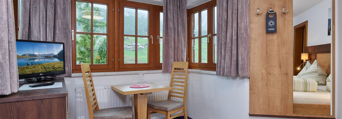 A cozy room with wooden decor and large windows. A table with chairs and a television are also available. - Haus Fontana - Sölden