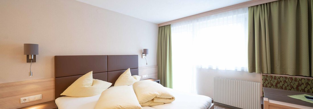 A modern bedroom with a large double bed and beige walls. The windows are decorated with green curtains and let in plenty of light. - Haus Fridolin - Sölden