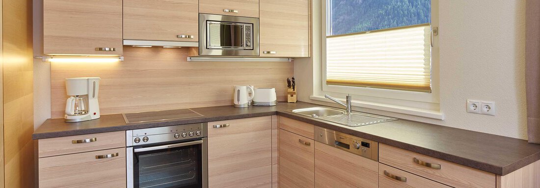 A modern kitchen with light wood cabinets and functional equipment. It features a stove, a microwave, and plenty of daylight from the window. - Haus Fridolin - Sölden