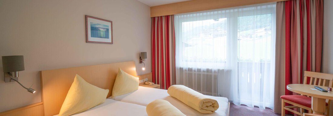 A cozy hotel room with two beds, yellow pillows, and a small table. Large windows let in plenty of daylight. - Haus Fridolin - Sölden