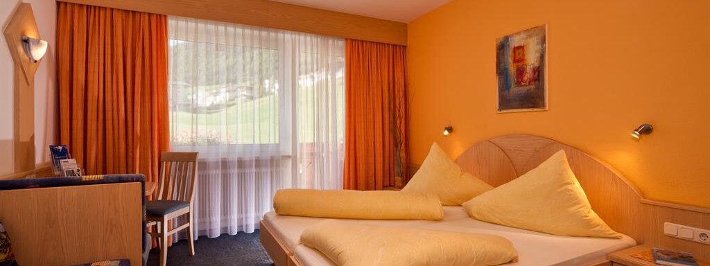 Zimmer A bright bedroom with orange walls and large windows. There is a double bed with large pillows and a cozy armchair. - Haus Fridolin - Sölden