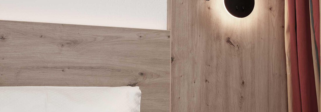 A modern wall lamp on a wood panel next to a bed. The lamp emits warm light and creates a cozy atmosphere. - Haus Gabriela - Sölden