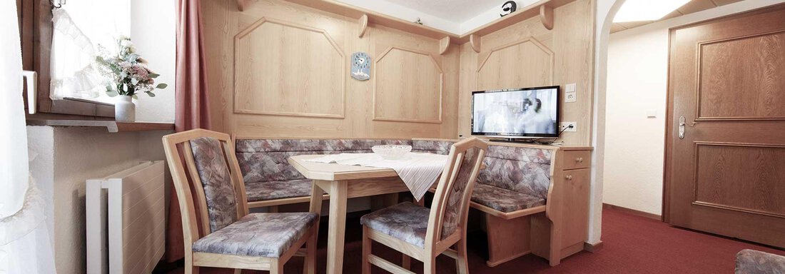 Appartement-01B | © Falkner Georg (CC C) A cozy dining area with wooden paneling and a table. In the background, there is a television and a window with curtains visible. - Haus Gabriela - Sölden