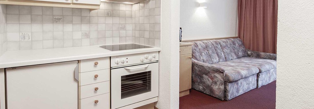 A bright kitchen with modern appliances and a seating area. The room has carpet flooring and a large window front with curtains. - Haus Gabriela - Sölden
