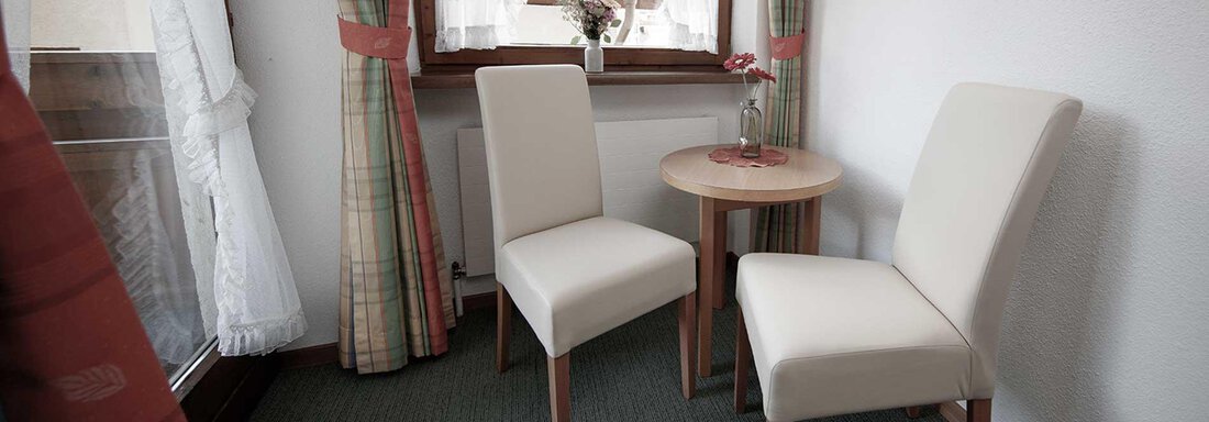 A bright room with two white chairs and a small round table. Through the window, you can see a friendly atmosphere with curtains and a small floral decoration. - Haus Gabriela - Sölden