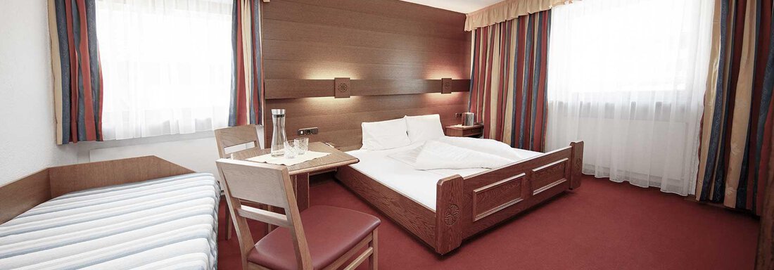 A simple hotel room with a double bed and a single bed. The room has large windows and a warm tone. - Haus Gabriela - Sölden