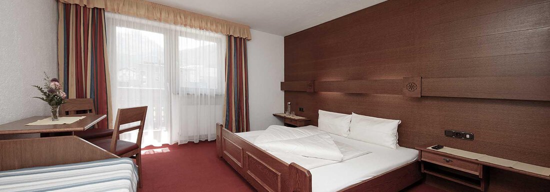 A simple hotel room with a double bed and a table with a chair. The room has large windows that let in a lot of light. - Haus Gabriela - Sölden
