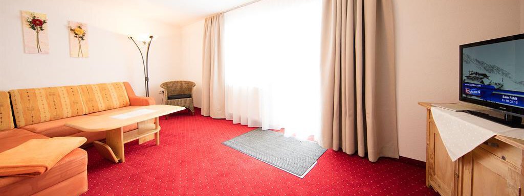 A cozy living room with a red carpet and a large window front. There is a sofa, a chair, and a television. - Haus Gondelblick - Sölden