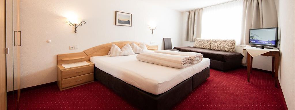 A cozy hotel room with a red carpet and a large window front. There is a double bed, a seating area, and a television. - Haus Gondelblick - Sölden