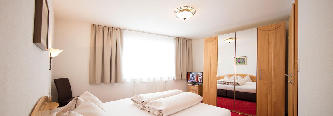 A cozy bedroom with a large bed and a reddish carpet. The room has windows with curtains and a wardrobe. - Haus Gondelblick - Sölden