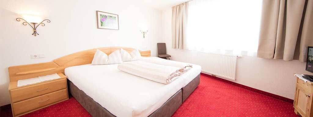 A cozy hotel room with a large double bed and red-spotted carpet. The room has plenty of daylight thanks to the large window. - Haus Gondelblick - Sölden
