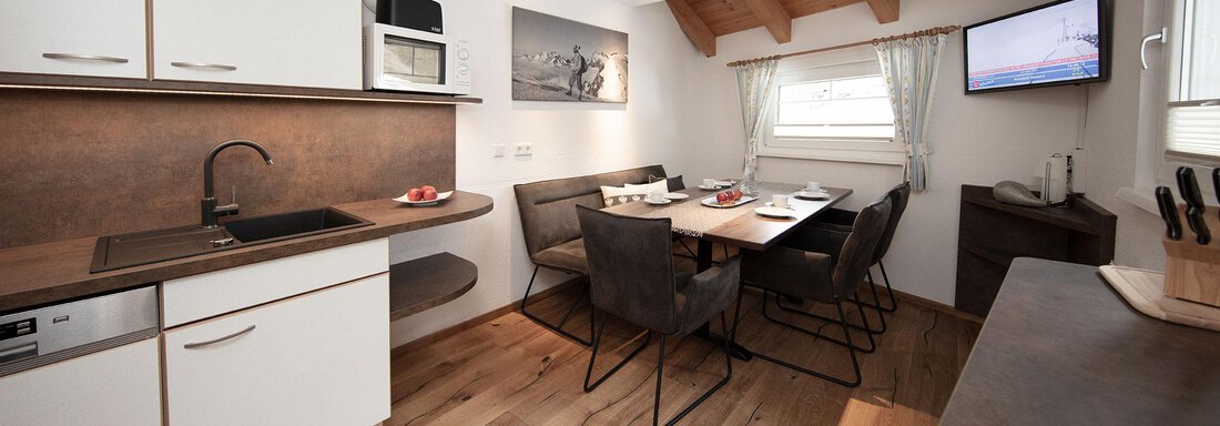 A modern kitchen with a dining area and wooden floor. Light pours in through large windows, creating a bright atmosphere. - Haus Gondelblick - Sölden