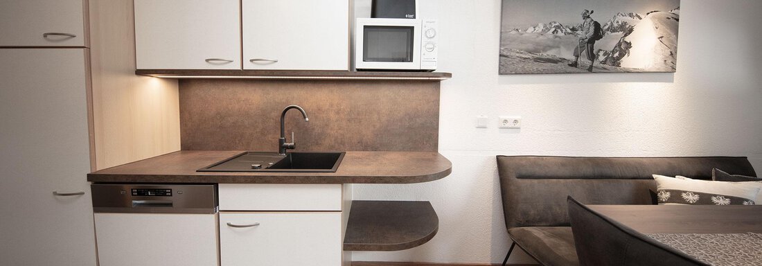 A modern kitchen with white cabinets and a wooden work area. In the background, there is a cozy seating area and a piece of art on the wall. - Haus Gondelblick - Sölden