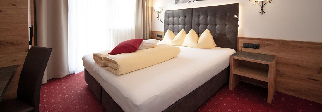 A modern bedroom with a large bed and bright pillows. The walls are made of wood, and there is a window with curtains. - Haus Gondelblick - Sölden