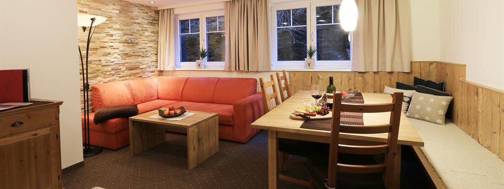 A modern living room with a red sofa and a wooden table. The room is bright and cozy with large windows and a dining area. - Haus Gondelblick - Sölden