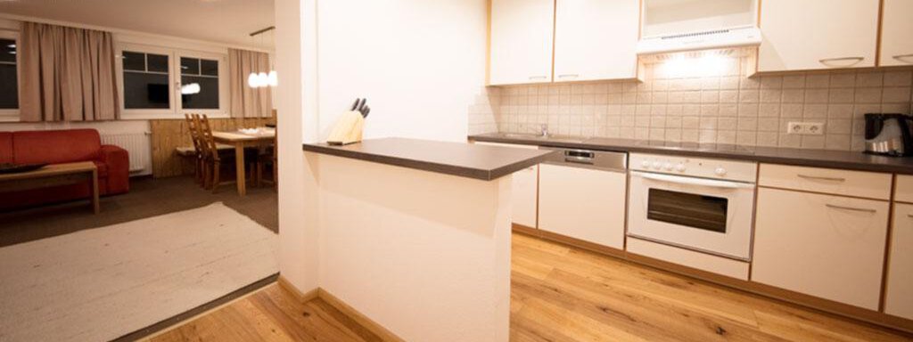 A modern kitchen with bright cabinets and a large wooden floor. In the background, a cozy living area is visible. - Haus Gondelblick - Sölden
