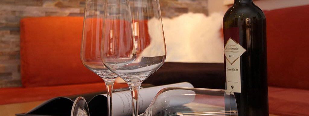 A table with three wine glasses and a bottle of red wine. In the background, there is a cozy seating area with a warm color tone. - Haus Gondelblick - Sölden