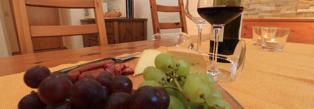 A table set with a plate of grapes, sausage, and cheese. In the background, there is a glass of red wine and a television. - Haus Gondelblick - Sölden