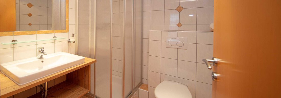 A modern bathroom with a shower, sink, and toilet. The floor is tiled and there is a large mirror above the sink. - Haus Gondelblick - Sölden