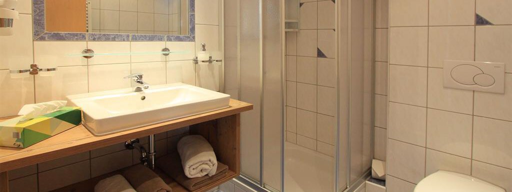 A modern bathroom with a shower, sink, and toilet. It features bright tiles and wooden shelves with towels. - Haus Gondelblick - Sölden