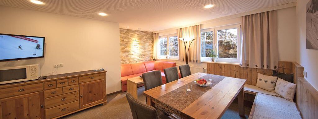 A cozy living room with a large dining table and wooden details. There is a sofa, a television, and windows with plenty of daylight. - Haus Gondelblick - Sölden
