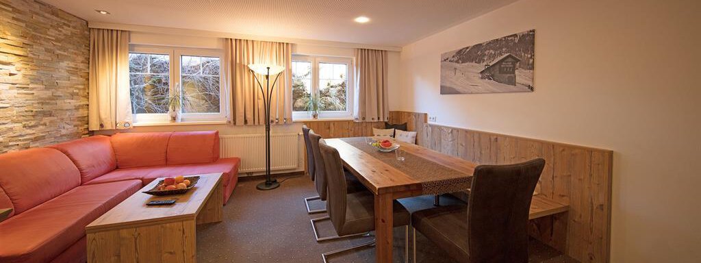 A modern living room with a red couch and a large wooden dining table. The rooms are bright and inviting, with large windows and soft lighting. - Haus Gondelblick - Sölden