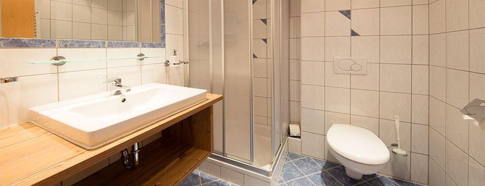 A modern bathroom with a shower and a sink on a wooden shelf. The walls are tiled with light tiles, and the floor is blue. - Haus Gondelblick - Sölden