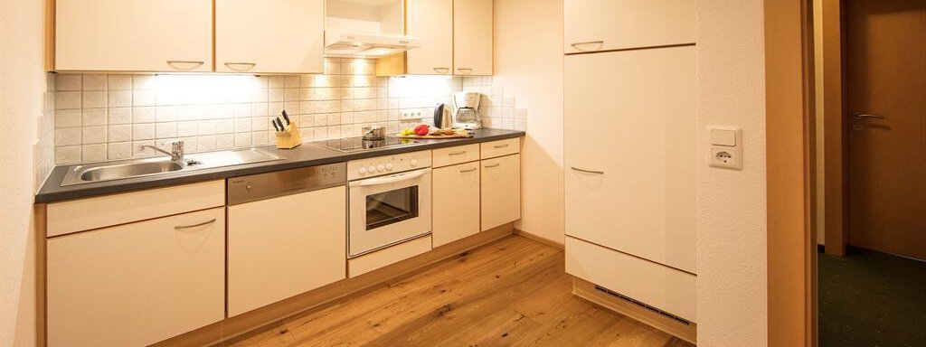 A modern kitchen with wooden-colored flooring and white cabinets. The kitchen features a refrigerator, a stove, and countertops. - Haus Gondelblick - Sölden